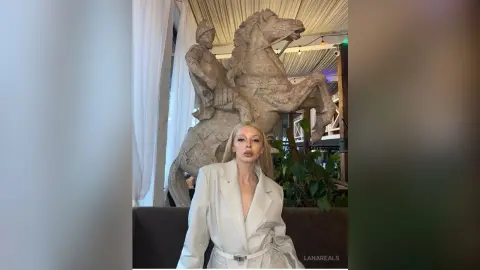 Elegant blonde woman in a white blazer sitting before a large stone equestrian statue in a bright indoor setting.
