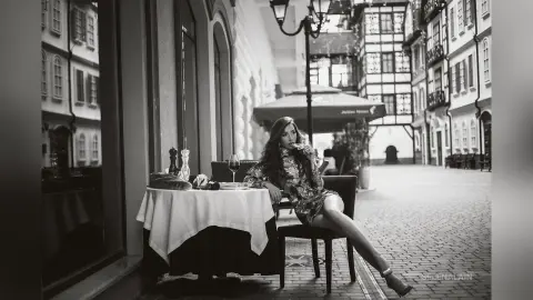 Elegant woman in a floral dress sitting at an outdoor cafe table in a charming European cobblestone street.