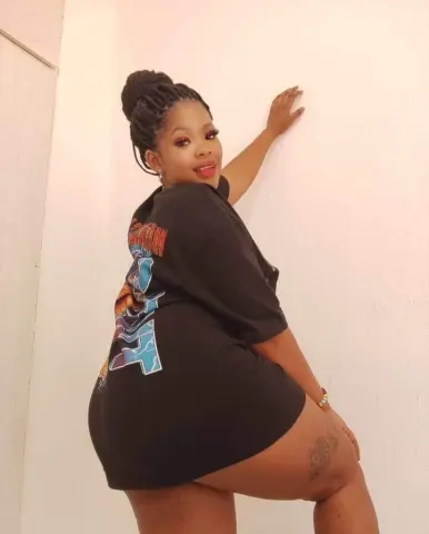 Smiling Black woman in a black graphic oversized t-shirt dress posing against a white wall.