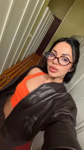 Young woman in glasses and a brown leather jacket over an orange top posing in a modern indoor setting.