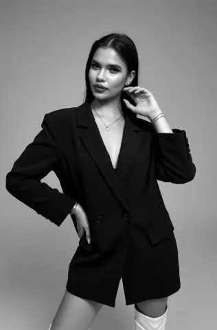 Elegant woman in a black oversized blazer and necklace posing against a minimalist grey studio background.