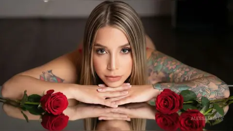 Blonde woman with colorful arm tattoos lying on a reflective surface with red roses in a moody setting.