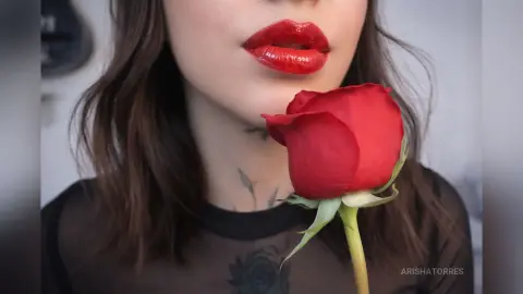 Woman with red lipstick and neck tattoos holding a single red rose in a moody, romantic setting.