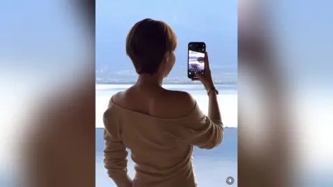 Woman in beige off-the-shoulder sweater taking a photo of a serene ocean view from a balcony.