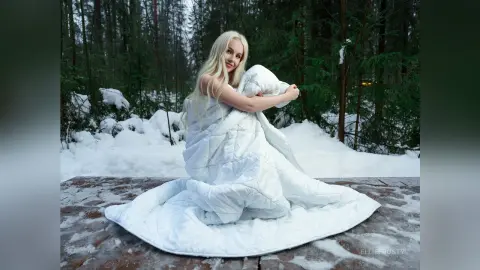 EllieFrosty sits on a wooden platform in a snowy forest, wrapped in a large white duvet that covers her body.