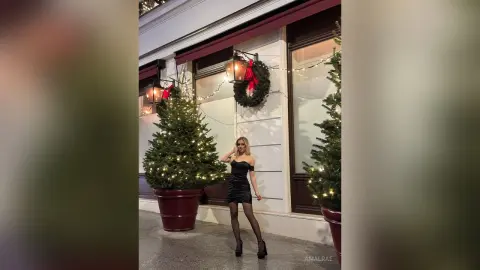 Elegant woman in a black cocktail dress posing by festive Christmas trees in a chic urban setting.