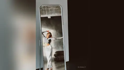 Young woman in a grey long-sleeve crop top and white trousers posing in a mirror with moody lighting.