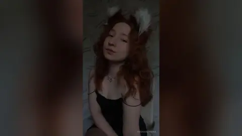 MadgeRemerez poses with long red hair and white furry ears, wearing a black camisole and a heart necklace.