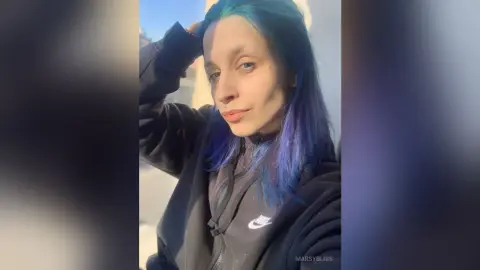 Young woman with blue hair wearing a black Nike hoodie in bright outdoor sunlight.