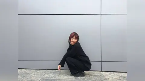 Young woman in a black oversized sweater and pants sitting against a minimalist grey paneled wall.