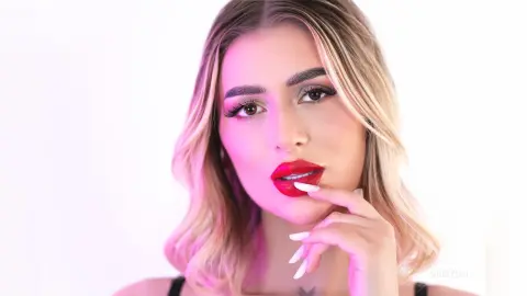 DaisyLin poses with her hand near her red lips, showing her blonde hair and light makeup against a white background.
