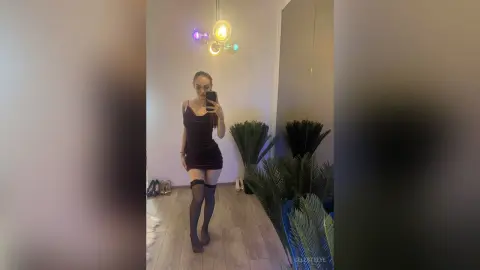 Young woman in a dark velvet mini dress and black thigh-high stockings posing in a modern minimalist room.