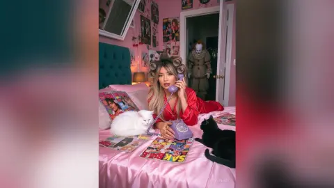 Blonde woman in red silk pajamas using a vintage purple phone on a pink bed surrounded by magazines and cats.
