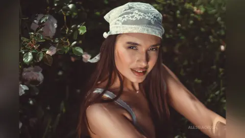 AlessiaCrush wearing a white lace headscarf and a blue bra while posing outdoors near pink roses.
