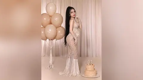 Elegant woman in a white lace gown celebrating a 23rd birthday with beige balloons and a cake in a dreamy setting.