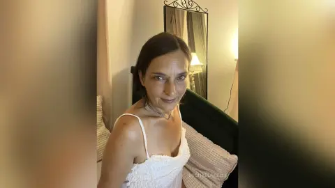 MichaelaBelle looks at the camera wearing a white spaghetti strap top with her dark hair down.