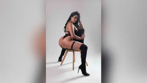 Curvy woman in black lingerie and over-the-knee boots posing on a chair against a white studio background.