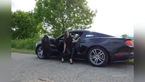 Stylish woman in black outfit stepping out of a sleek black sports car on a sunny roadside.
