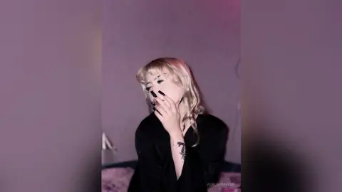 Blonde woman in a black outfit posing thoughtfully in a moody, dimly lit room.