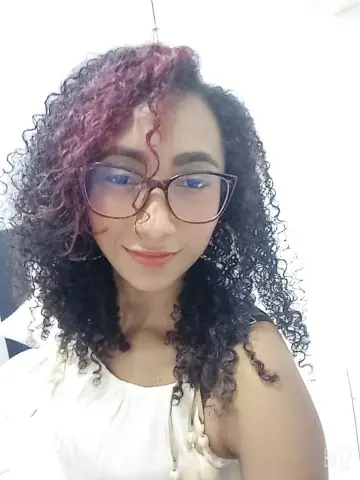 Smiling woman with curly dark hair and glasses wearing a white sleeveless top in a bright indoor setting.