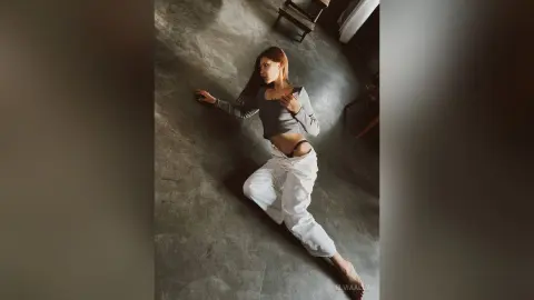 Fashionable woman in a grey crop top and white trousers sitting on a concrete floor in a moody indoor setting.