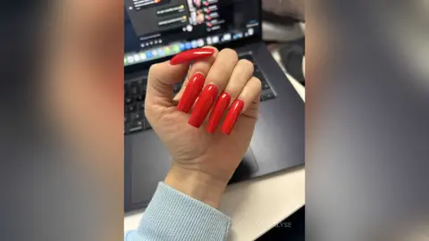 Close-up of long red almond-shaped manicured nails held against a laptop in a cozy home office setting.