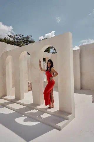 Elegant woman in a red evening gown posing in a minimalist white arched courtyard under a bright sunny sky.