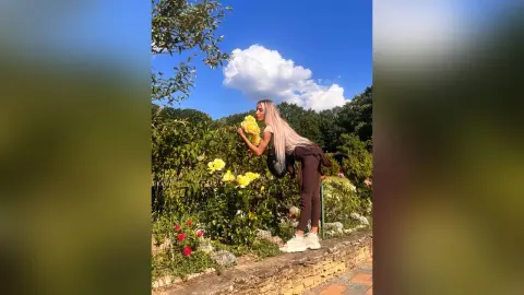 Blonde woman in casual dark outfit smelling yellow roses in a sunny summer garden.