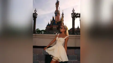 NickyAshcroft poses in a white sundress and sunglasses in front of a large castle, showing off her blonde hair and slim figure.