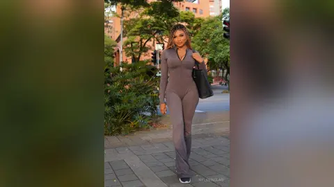 Stylish woman in a brown long-sleeve jumpsuit standing on a city sidewalk near lush green bushes.