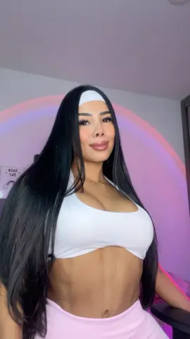 Smiling woman with long black hair in white sports bra and pink skirt posing in a room with pink neon lighting.