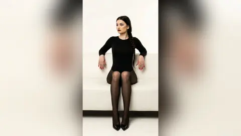 ZenyaSin sits on a white couch wearing a black long-sleeve bodysuit and sheer black tights with black heels, her long dark hair styled in a braid.