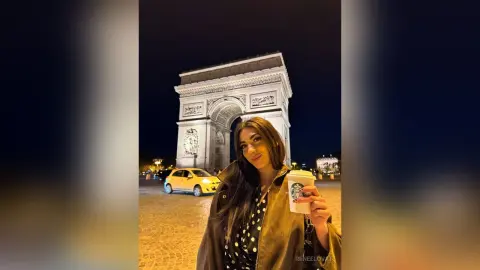 Stylish woman in a dark jacket holding a coffee cup in front of the illuminated Arc de Triomphe at night.