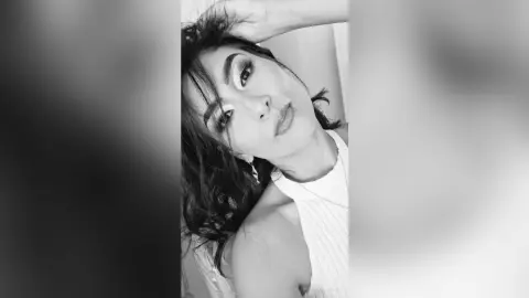 MaddyBrohms poses with her hand in her dark hair, wearing a white tank top in this black and white close-up shot.