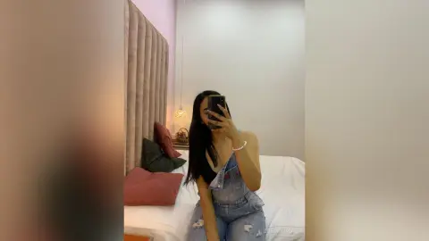 Young woman in denim overalls taking a mirror selfie in a cozy, modern bedroom with soft lighting.