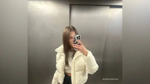 Young woman in a white puffer jacket taking a mirror selfie in a modern elevator.