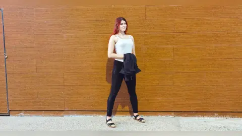 Red-haired woman in a white tank top and black leggings standing against a wooden wall.