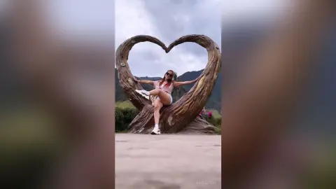 Young woman in a pink tank top sitting in a heart-shaped wooden sculpture amidst a scenic mountain landscape.
