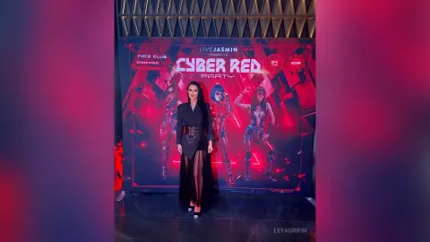 LeyaGrifin stands in front of a Cyber Red Party backdrop wearing a black outfit with a wide belt.