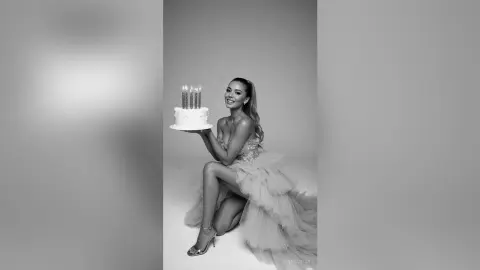 SerenaStar smiles while holding a lit birthday cake, wearing a light-colored ruffled tulle gown and high heels.