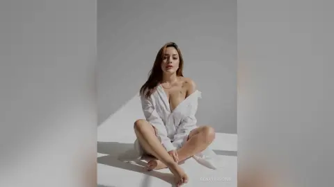 Young woman in a white oversized shirt sitting cross-legged in a bright, minimalist studio setting.