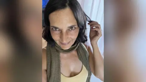 MichaelaBelle smiles while tucking a strand of her dark hair behind her ear, wearing a tan tank top and an olive green scarf.