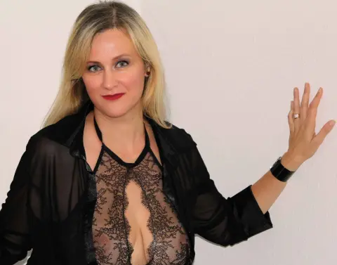 MarleneBloem poses in a black sheer lace top and open black shirt, waving her hand against a plain white wall.