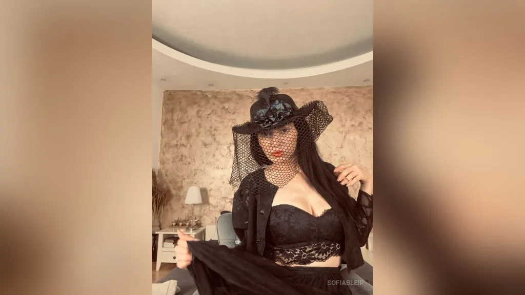 SofiaBleir — SofiaBleir wears a black lace outfit and a wide-brimmed veiled hat while posing in a room.