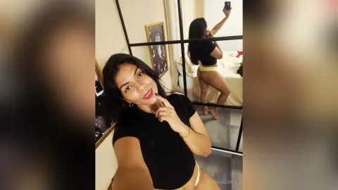 Smiling woman in a black t-shirt taking a mirror selfie in a bright bedroom setting.