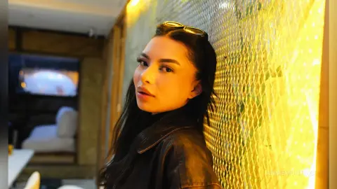 Young woman in a black leather jacket leaning against a glowing gold textured wall in a moody indoor setting.
