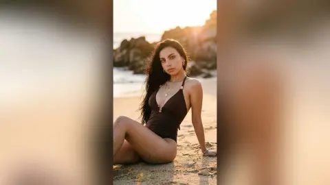 Young woman in a dark brown one-piece swimsuit sitting on a sandy beach during a golden sunset.