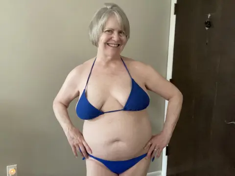 Smiling mature woman in a blue string bikini standing in a brightly lit room.