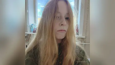 Young woman with long blonde hair wearing a dark green top in a bright indoor setting.