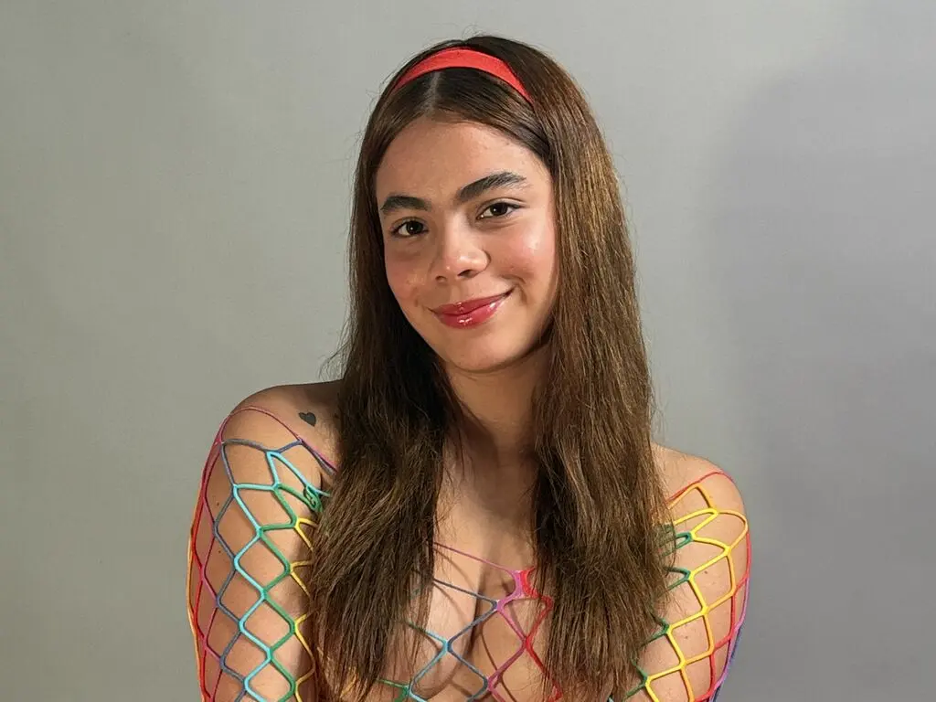 Smiling young woman with long brown hair wearing a colorful rainbow fishnet mesh top against a grey background.